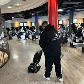 Eine Reinigungskraft bearbeitet mit einer Maschine den glänzenden Steinboden in einem Fitnessstudio mit Cardio-Geräten im Hintergrund.