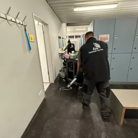 Zwei Mitarbeiter reinigen einen Umkleidebereich mit Spinden und Waschraumzugang, ausgestattet mit professionellem Reinigungswagen.