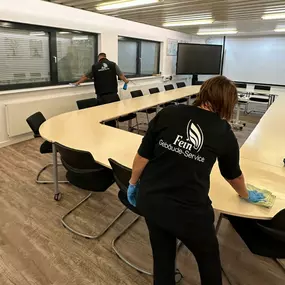 Ein Team von Fein Gebäude-Service wischt Oberflächen im Konferenzraum mit großem Besprechungstisch, moderner Technik und vielen Stühlen.