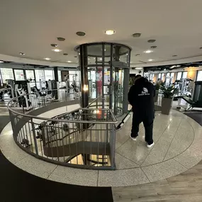 Eine Reinigungskraft säubert den Boden in einem gut ausgestatteten Fitnessstudio. Im Mittelpunkt steht ein Glasaufzug, umgeben von modernen Trainingsgeräten.