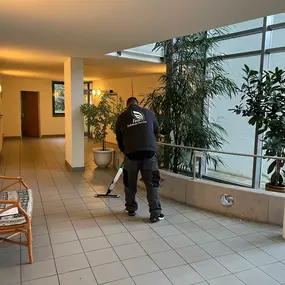 Eine Reinigungskraft bei der Arbeit in einem hellen, begrünten Flurbereich eines Gebäudes mit Sitzgelegenheiten.