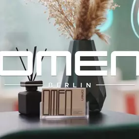Modernes Detail im OMEN Tattoo Studio Berlin: stilvolle Trockenblumen, Duftstäbchen und Visitenkarte vor Glasfront mit leuchtendem Logo. Designorientiertes Ambiente im Studio Berlin Mitte.
