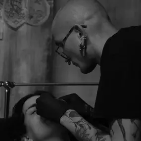 Piercer mit Brille und Tattoos arbeitet konzentriert an einem Gesichtspiercing einer Person im Studio.