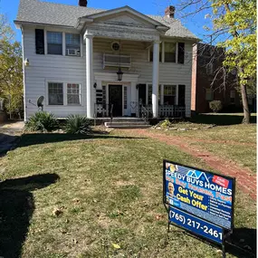 This image shows a classic two-story white house with traditional architecture, including large columns framing the front porch, located on a spacious lawn in a residential neighborhood. The property appears to require some maintenance or repairs, making it ideal for homeowners interested in selling quickly. Prominently displayed in the front yard is a bold and clear sign for Speedy Buys Homes, advertising quick home purchases for cash, with highlighted services including buying properties in an