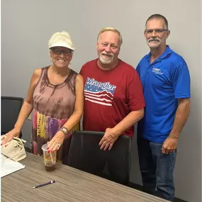 This image captures a joyful moment featuring Tim from Speedy Buys Homes with happy homeowners, Mike and Tammy, celebrating the successful sale of their property. Tim is seen wearing a professional blue Speedy Buys Homes polo, reflecting the company's brand and trustworthiness. Mike and Tammy appear delighted, standing closely together, smiling warmly and clearly satisfied with their stress-free home-selling experience. The atmosphere suggests comfort, trust, and positive relationships, highligh
