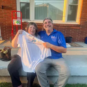 This image captures a joyful moment between Ryan from Speedy Buys Homes and a satisfied customer celebrating a successful property sale. Seated together on the front porch steps, both are smiling warmly and proudly holding up a branded Speedy Buys Homes t-shirt, highlighting the friendly and personal relationship between the company and its clients. Ryan, wearing a branded blue polo, represents professionalism and trust, while the homeowner's happiness reflects the positive experience provided b