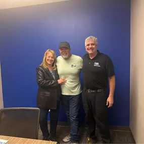 This image features Ryan from Speedy Buys Homes standing happily alongside the Lowe family after successfully closing on their home sale. They are smiling warmly in a comfortable and professional office setting with a vibrant blue background, symbolizing trust and confidence. Ryan, dressed professionally in a black branded polo, embodies the company's commitment to approachable, trustworthy service. The Lowe family's smiles and relaxed poses reflect their positive experience and satisfaction wit