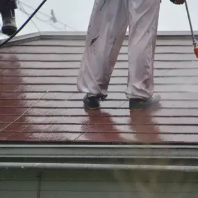 A person in work attire and boots is pressure washing a red-brown shingle roof. Water sprays from the wand, cleaning the roofing material. Another person's legs are visible in the top left corner.