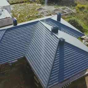 An aerial view shows a house with a dark gray metal roof featuring multiple gables and chimneys. The roof's texture is visible, and the surrounding area includes a lawn, trees, and another building under construction.
