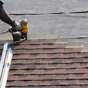 A person wearing blue jeans and work gloves is using a nail gun to install brown and gray asphalt shingles on a roof. Part of a silver ladder is visible on the left, and black underlayment is present on the roof's surface.