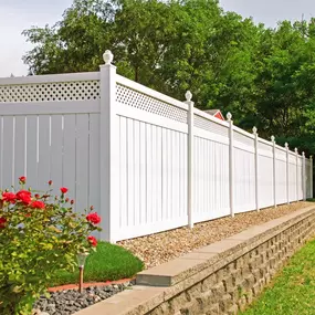 The image shows a long, white vinyl fence with decorative lattice on top, running along a stone retaining wall. Red flowers are visible in front of the fence, and green trees form a backdrop.