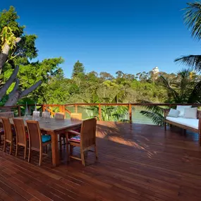 A spacious wooden deck features an outdoor dining table with chairs and a cushioned bench. The deck overlooks lush greenery and a body of water under a clear blue sky, suggesting a scenic waterfront setting for outdoor entertaining.