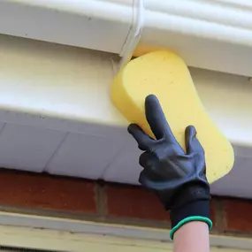 A gloved hand cleans a white gutter with a yellow sponge. The gutter is attached to a white structure above a red brick wall. The hand wears a black glove with a green trim.