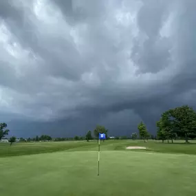 Dramatic summer storm clouds over the green at Audubon Golf Course, with a blue flag and bunker in the background.