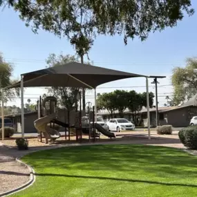 A playground with a slide and a canopy is surrounded by a grassy area and a sidewalk.