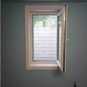 This interior photo showcases a premium egress window installation by Egress of Long Island, featuring a high-quality casement window with white vinyl framing. The window is professionally installed in a newly finished basement wall with a soft blue-green paint color. The window is shown in the open position, demonstrating its full functionality for emergency escape purposes. Through the window, you can see the corrugated egress well walls that provide structural support while allowing natural l