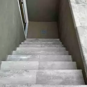 This image depicts a full-depth concrete basement walkout installation by Egress of Long Island, showcasing their mastery of large-scale basement solutions. The photo captures a view from above, revealing professionally poured concrete stairs descending to a code-compliant door at the bottom. The stairs feature precision-cut treads with proper depth and height measurements to ensure safe passage during emergencies. The smooth, sealed concrete walls provide excellent water resistance while creati