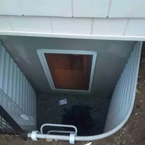 This image showcases a professionally installed egress window well by Egress of Long Island, demonstrating superior construction techniques that exceed local code requirements. The photo captures a curved, galvanized steel egress well designed for maximum durability and structural integrity. The well features a premium heavy-duty egress window with a protective screen covering, allowing for both natural light penetration and secure ventilation. The floor of the well is properly prepared with a w