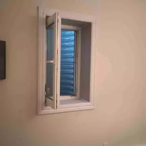 This image showcases a completed egress window installation in a basement by Egress of Long Island. The professionally installed egress features a modern white casement window with clear glass panes and operational hardware, allowing it to fully open for emergency exit. The window is framed with clean white trim that complements the neutral wall color, creating a seamless integration with the interior design. Beyond the window, a white egress well is visible, which meets local building codes for