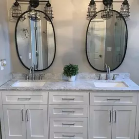 This bright bathroom features a double vanity with white cabinets, a marble countertop, and two oval mirrors. Industrial-style cage light fixtures illuminate the space, complemented by a small green plant.