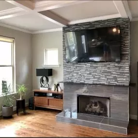 This inviting living room features a modern fireplace with a grey tiled surround and ledger stone accent wall, topped by a mounted television. Hardwood floors and white coffered ceilings complete the bright space.