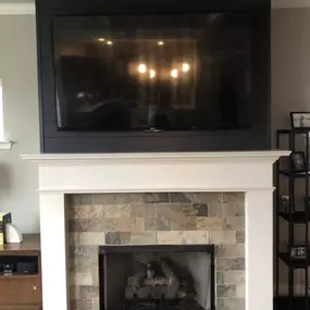 This close-up shows a cozy fireplace with a multi-toned stone tile surround and a white mantel. Above it, a large TV is mounted on a dark shiplap accent wall, reflecting interior lights.