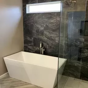 This luxurious bathroom features a modern white freestanding tub set against a dramatic dark, veined tile accent wall. Light wood-look plank tiles are arranged in a herringbone pattern on the floor, adding warmth to the space.