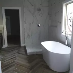 This modern bathroom features a stylish wood-look herringbone tile floor. A freestanding white bathtub is positioned near a window, opposite a shower with large format white marble-look tiles, currently undergoing installation.