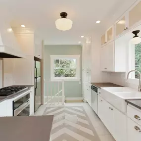 A beautifully remodeled all-white galley kitchen features a stove, oven, farmhouse sink, and elegant gray countertops. Herringbone pattern floor tiles lead towards a window with a staircase.