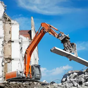 A large orange excavator is demolishing a white concrete building under a clear blue sky. Debris and rubble are scattered at the base of the partially collapsed structure.