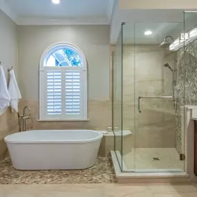 A modern bathroom features a glass-enclosed shower, a separate white bathtub, and brown wood cabinets. Towels hang neatly beside a window with white shutters.