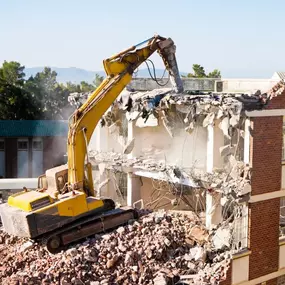 A large yellow excavator with a hydraulic breaker attachment is actively demolishing a brick building, leaving a trail of rubble and dust, under a clear sky.