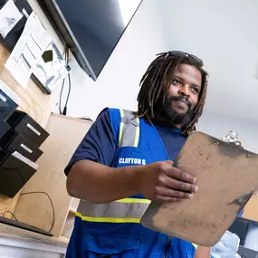 A Builders FirstSource team member holds a clipboard inside a dispatch or logistics office. He wears a reflective safety vest over a navy shirt and stands in front of a wall-mounted document organizer and computer station.