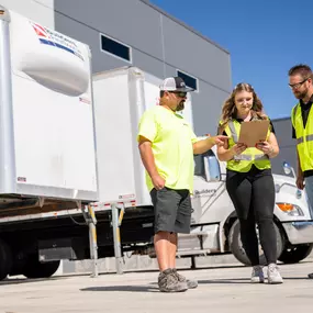 Three Builders FirstSource team members in safety vests review paperwork on a clipboard outside a distribution center, collaborating in front of delivery trucks under a clear blue sky.
