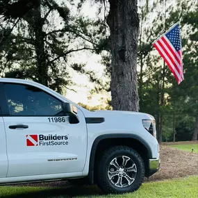 A white Builders FirstSource company truck parked on a grassy area beside a large pine tree. An American flag is mounted on the tree, gently waving in the breeze. The scene captures a sense of patriotism and pride in service, set against a backdrop of tall trees and a soft, evening sky.