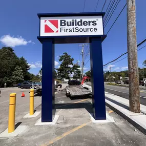Street sign for Builders FirstSource outside a construction supply store, located next to a busy road under a clear blue sky.