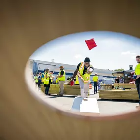 A group of Builders FirstSource employees in neon safety vests play a game of cornhole outdoors. The image is captured through the cornhole board’s hole, focusing on a teammate in mid-throw with a red bean bag. Others look on in a lively and fun team-building atmosphere.