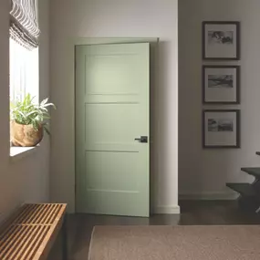 A cozy, modern hallway with a soft green, three-panel interior door slightly ajar. The space includes a woven bench, a window with a potted plant, a natural fiber rug, and a wall adorned with three black-and-white framed photographs. The design is minimal and welcoming.