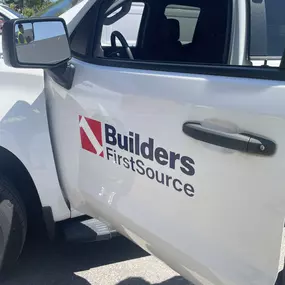 Side view of a white Builders FirstSource work truck with company logo on the door, parked outdoors on a sunny day.