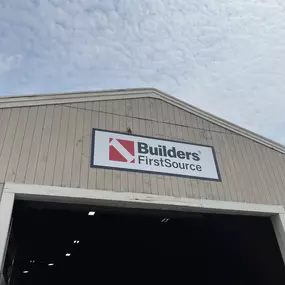 Exterior of a Builders FirstSource warehouse building with company sign mounted above the entrance, under a partly cloudy sky.