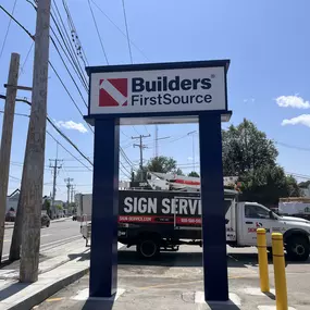 Street sign for Builders FirstSource outside a construction supply store, located next to a busy road under a clear blue sky.