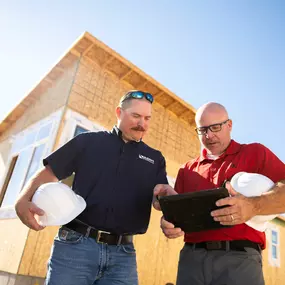 Two Builders FirstSource team members hold hard hats while reviewing construction plans on a tablet. They stand in front of a partially built house with wood framing under clear blue skies, representing planning, collaboration, and field operations.