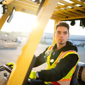 A Builders FirstSource team member operates a yellow forklift in a lumber yard during golden hour. He wears a black hoodie, neon yellow safety vest, and gloves, with safety goggles clipped to his chest. The background shows construction materials and a bright sun flare.