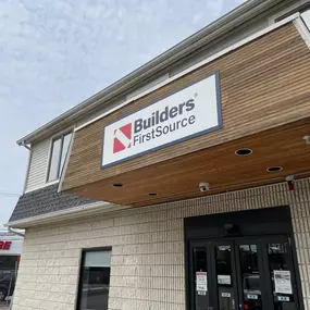 Front entrance of a Builders FirstSource retail building with company sign above the door, featuring a wood-accented overhang and brick exterior.
