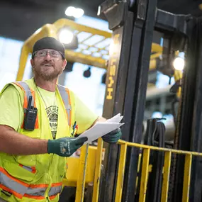 Builders FirstSource team member in a high-visibility vest smiling while holding paperwork next to a forklift inside a busy warehouse, reflecting professionalism and a commitment to service.