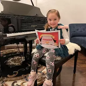 This photo captures a heartwarming and proud moment at a music studio. A young girl sits happily on a piano bench, holding a colorful “Wunderbies” achievement certificate with a bright smile on her face. Behind her, a sleek black Bergmann grand piano gleams, creating a beautiful and professional setting for her success.

The cozy space features an elegant rug, a soft faux-fur seat cushion, and a modern navy blue chaise lounge in the background, contributing to a warm and inspiring atmosphere. Na