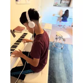 This photo captures a moment of focused concentration at Baney Jones Music Studio, where a young student is deeply engaged in piano practice. Wearing white Sony headphones, he is fully immersed in his music, concentrating on the keys of a sleek white digital piano. His posture and expression show dedication, discipline, and a love for learning.

In the background, another student is also practicing at a separate piano, reinforcing the studio’s engaging and structured learning environment. The wa