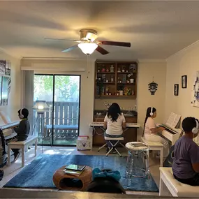 This photo shows a well-organized, cozy piano lesson studio where multiple students are engaged in their music practice. Each student is seated at their digital piano, wearing headphones, creating a focused and distraction-free environment for learning. The room is thoughtfully designed with soft lighting, modern decor, and artwork on the walls, including an artistic Eiffel Tower-themed gallery and a creative painting on the right side.

The studio’s open, inviting space includes a bright window