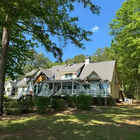 A large residential house with a new roof under construction, nestled amongst lush green trees. Several workers are visible on the rooftop, indicating active renovation or building.