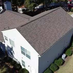 Aerial view of a suburban white brick building with a dark brown shingle roof, surrounded by trimmed bushes, trees, a driveway, and a well-maintained lawn under bright sunlight.